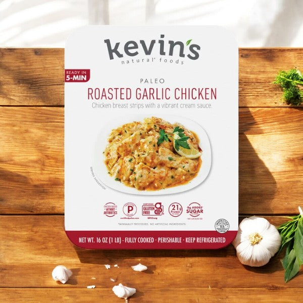 A package of Easy Lunches' Kevin's Natural Foods Roasted Garlic Chicken (1 Box - 16 oz) is displayed on a wooden surface, surrounded by garlic cloves and herbs. The pre-cooked meal states it's ready in just 5 minutes and boasts high-quality ingredients.