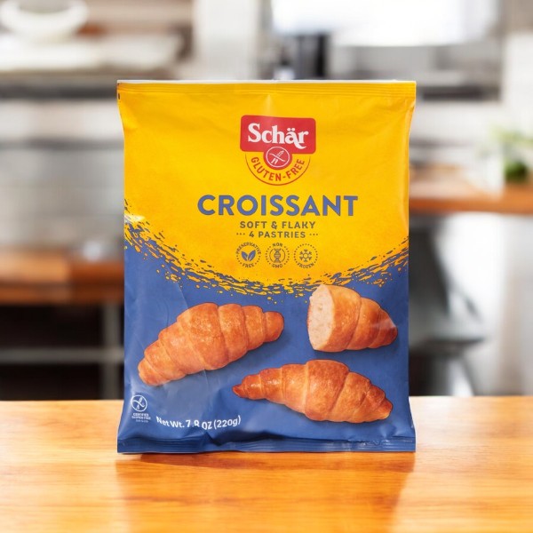 A package of Schar Plain Croissants, 7.8 oz - 1 Per Case, is displayed on a wooden surface, featuring an image of three light and flaky croissants alongside product information, emphasizing their versatile use for any meal.