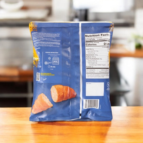 A blue bag of Schar Plain Croissants, 7.8 oz, sits on a wooden table, showcasing nutritional information and cooking instructions. Savor the light and flaky texture of these gluten-free croissants while discovering their versatility in both sweet and savory recipes.