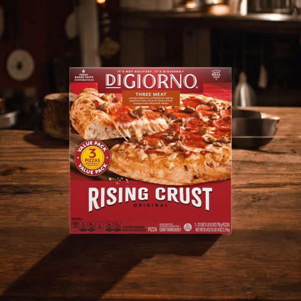 On a wooden counter lies a box of Easy Lunches DiGiorno Three Meat Pizza - 3 Count, 1 Pack, featuring an image of the cooked pizza generously topped with pepperoni, sausage, beef toppings, and mozzarella cheese. The packaging clearly states that it contains 3 pizzas.