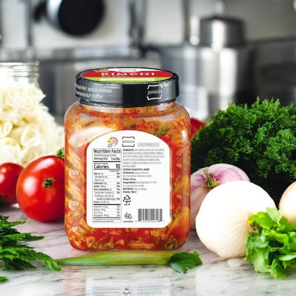 A jar labeled "Kimchi- Jongga Sliced Napa Cabbage Kimchi, 52.9 oz- 1 Pack" from Easy Lunches sits on a kitchen counter amidst fresh tomatoes, onions, greens, and herbs.