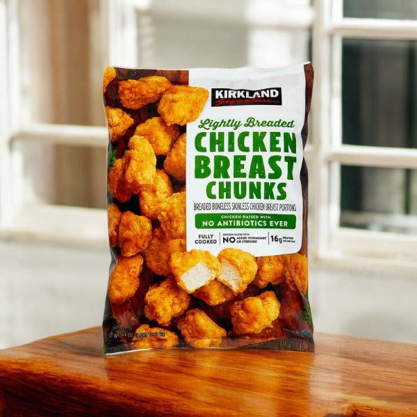 On a wooden surface, a bag of Kirkland Signature Lightly Breaded Chicken Breast Chunks, weighing 4 lbs., is illuminated by natural light streaming in from a nearby window. The package prominently features labels like "No Antibiotics Ever" and "Fully Cooked," providing reassurance to all poultry enthusiasts who might also appreciate premium seafood options such as shrimp.