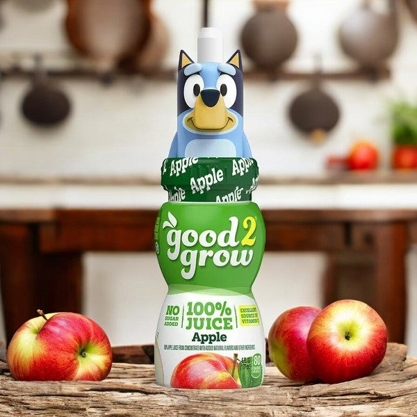 A 6 oz single-serve bottle of Easy Lunches' Good2grow Apple Juice, featuring 100% real apple juice and a collectible character top (character tops may vary). The background includes a wooden table adorned with various apples.