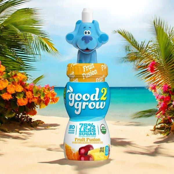 A bottle of Easy Lunches Good2grow Fruit Fusion Juice Single Serve, 6 oz - 1 Count (Character Tops Vary), with 75% less sugar and a blue dog character topper, featuring 100% real fruit juice, stands on a sandy beach with vibrant flowers and a tropical ocean in the background.