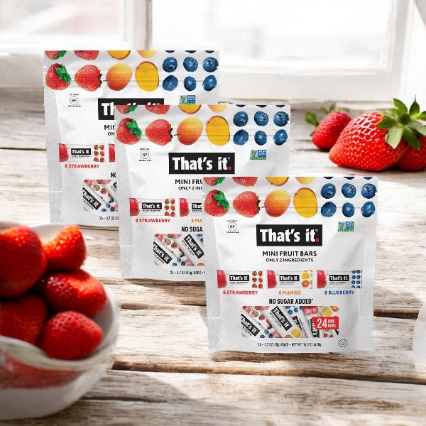 Displayed on a white surface with backlighting through a window, three packs of Easy Lunches "That's it" Mini Fruit Bars in strawberry, mango, and blueberry flavors sit alongside fresh strawberries.