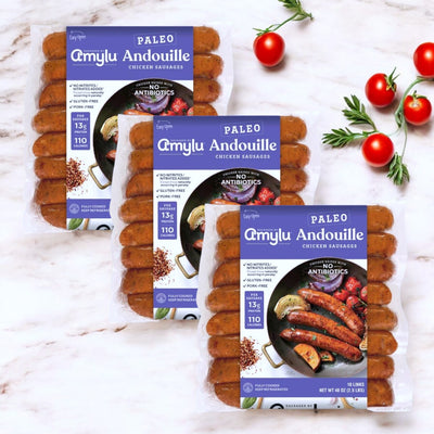 Three packs of Amylu Paleo Chicken Andouille Sausage – 16 Count (40 oz) are arranged on a light wooden surface, surrounded by cherry tomatoes and herbs. These sausages are gluten-free and feature bold Cajun flavor.