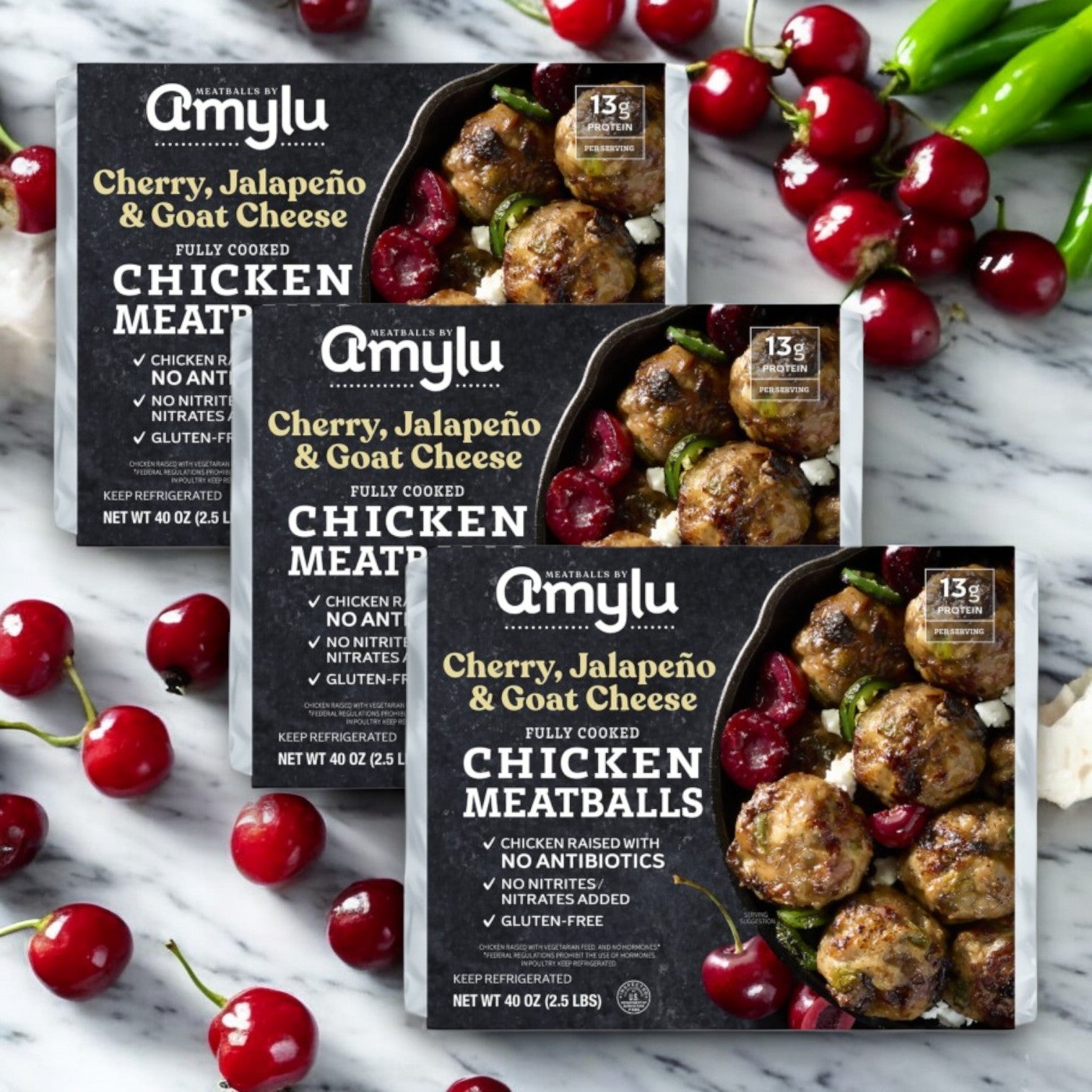Three packs of Easy Lunches Amylu Cherry, Jalapeno, and Goat Cheese Chicken Meatballs (40 oz) are displayed on a marble surface with fresh cherries and jalapeños scattered around.