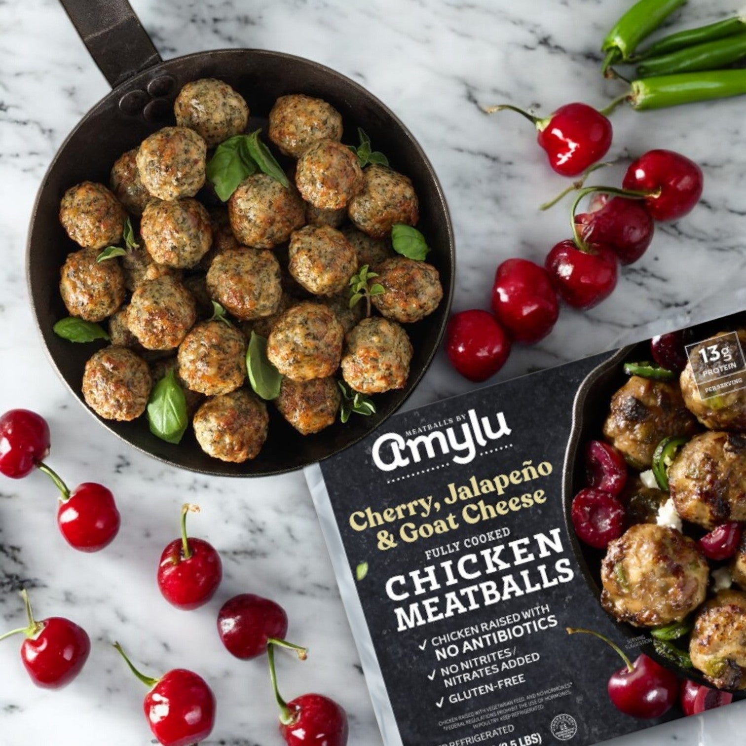 A skillet of chicken meatballs is placed beside fresh cherries, jalapeños, and a package labeled "Easy Lunches Amylu Cherry, Jalapeno, and Goat Cheese Chicken Meatballs, 40 oz, 3 Packs.