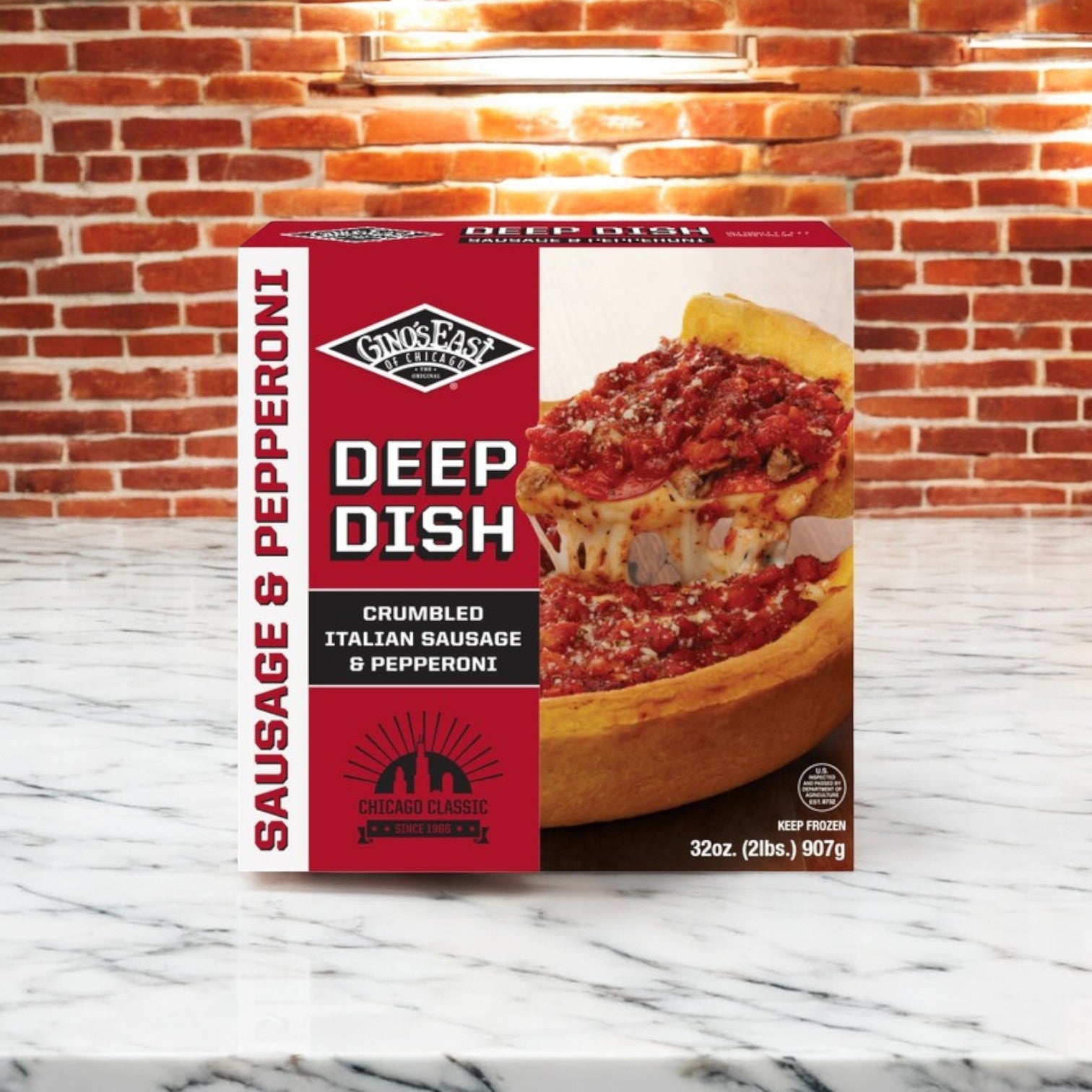 A Gino's East Pizza box from the 2 Pack Combo rests on a marble surface against a brick wall, epitomizing authentic Chicago-style with crumbled Italian sausage and pepperoni.