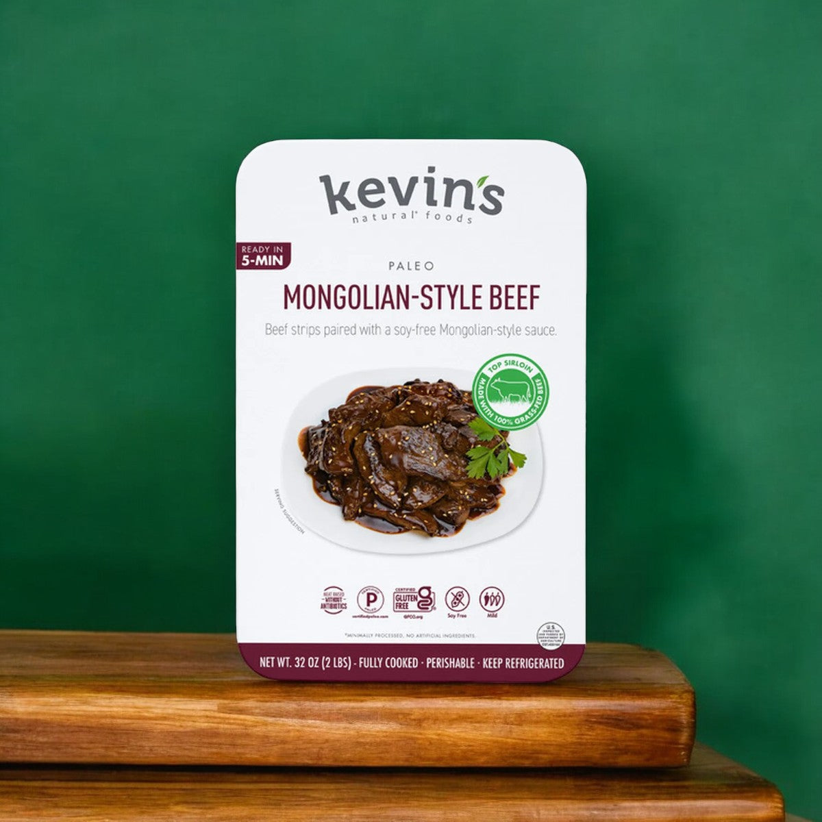 Kevin's Mongolian Beef, a 32 oz package from Kevin's brand, features gluten-free beef strips in a flavorful Mongolian-style sauce. The text highlights that it is paleo, soy-free, and ready in just 5 minutes. The product is presented against a green background on a wooden surface.