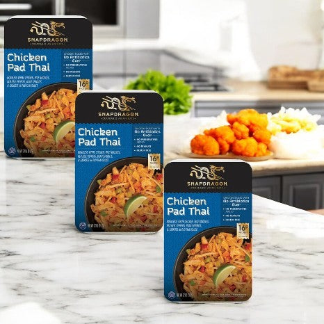 On a marble countertop, three boxes of Snapdragon Chicken Pad Thai, 32 oz-3 Count, capture the authentic restaurant-style allure with a backdrop of fried shrimp and colorful vegetables.