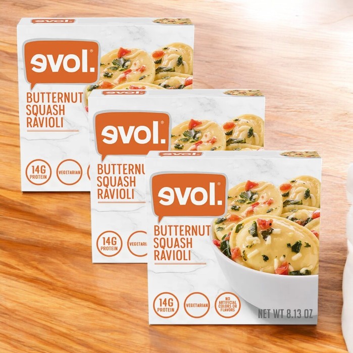 Three boxes of Evol Vegetarian Butternut Squash & Sage Ravioli, 8.13 oz each, are displayed on a wooden surface. The packaging emphasizes its 14g protein content and vegetarian label, highlighting the delicious flavor combination.