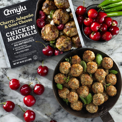 A skillet of tasty chicken meatballs with goat cheese sits next to a package of Amylu Cherry, Jalapeno, and Goat Cheese Chicken Meatballs, 40 oz, surrounded by fresh cherries and green chili peppers.