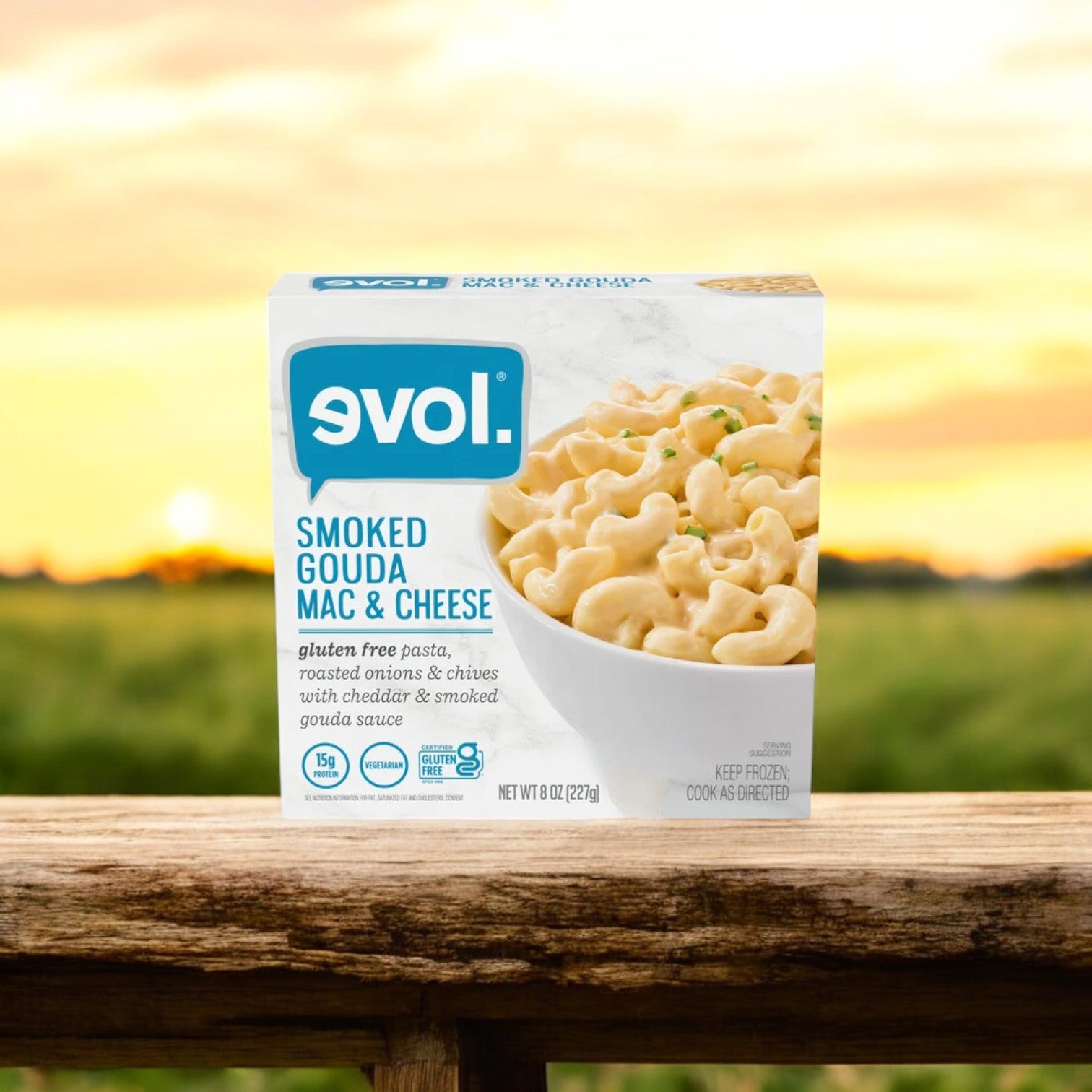 An 8 oz. package of Evol Gluten Free Smoked Gouda Mac and Cheese, a frozen meal renowned for its creamy smoked gouda cheese sauce, rests on a wooden railing against a blurred outdoor backdrop, inviting with its tempting flavors.