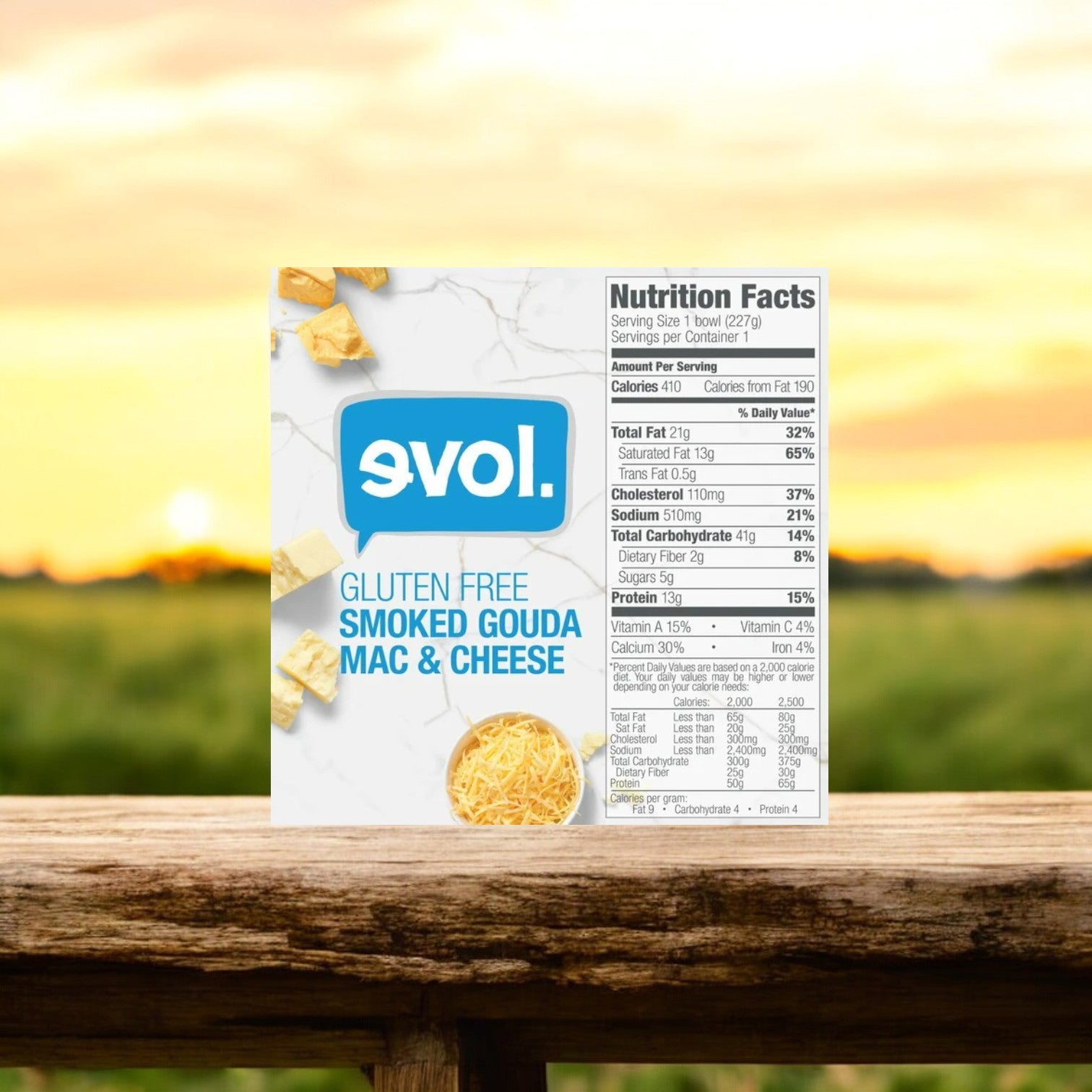 An 8 oz. frozen meal of Evol Gluten Free Smoked Gouda Mac and Cheese is elegantly placed on a wooden surface, boasting its gluten-free pasta and creamy smoked gouda cheese sauce that capture attention, with its nutritional facts prominently displayed on the side against a softly blurred outdoor background.