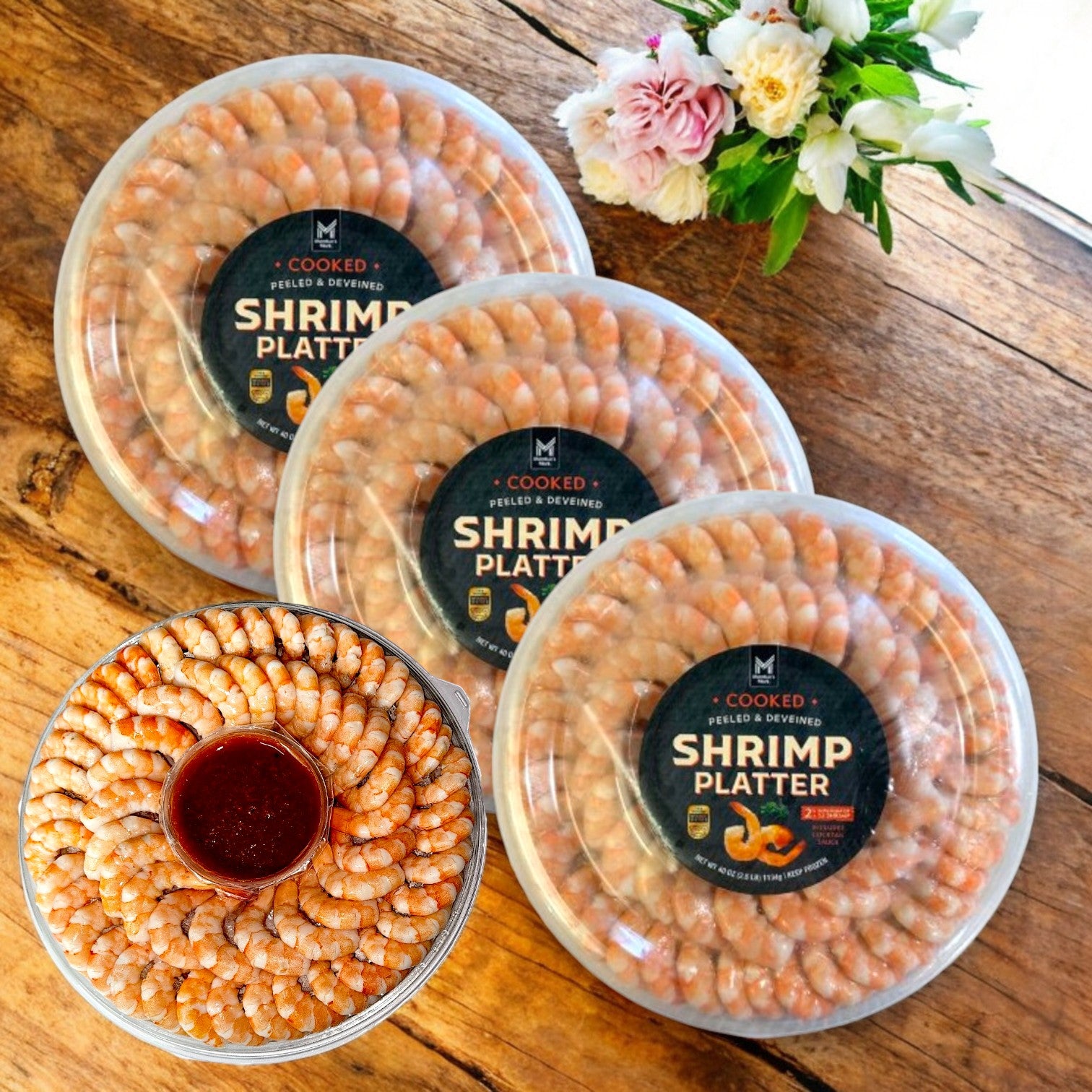 Three Member's Mark Shrimp Platter packs with cocktail sauce (7.5 lbs total) are elegantly packaged on a wooden table, alongside one open platter and flowers in the background.