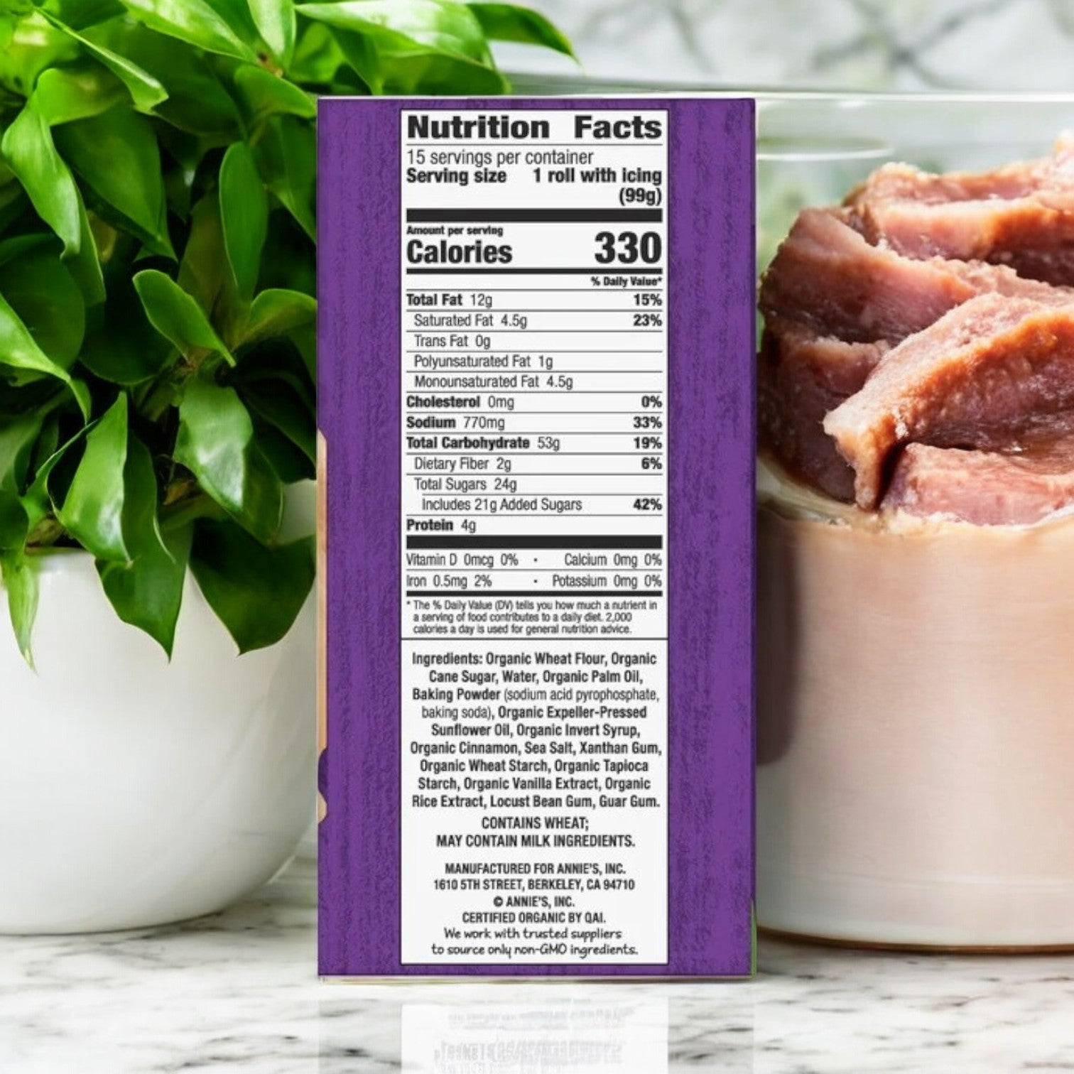 The box of Annie's Organic Cinnamon Rolls, 17.5 oz, 3-count from Annie's brand, displaying comprehensive nutrition facts and an ingredient list featuring natural ingredients, is positioned beside a potted plant and meat slices in the background.