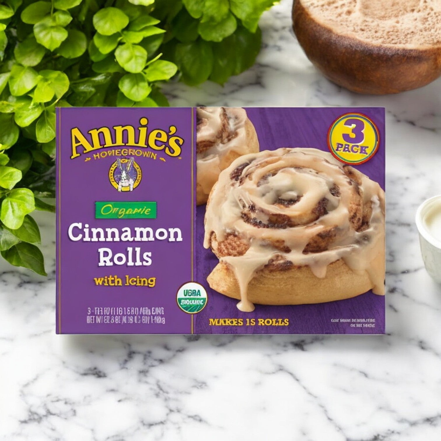 A box of Annie's Organic Cinnamon Rolls rests on a marble countertop beside a potted plant. With natural ingredients, the packaging showcases a 3-count box weighing 17.5 oz and contains a total of 15 delectable rolls complete with creamy icing.