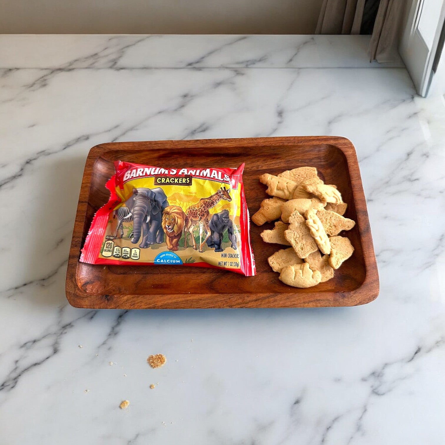 A packet of Barnum's Original Animal Crackers (1 oz- 1 Count) sits on a wooden tray with several animal-shaped crackers scattered around, all displayed on a marble surface—perfect for anyone seeking calcium snacks.