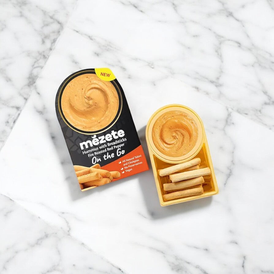 A Mezete Fire Roasted Red Pepper Hummus with Breadsticks On The Go snack pack is placed on a marble surface. The pack is open, revealing the creamy hummus made from natural ingredients and crunchy breadsticks inside.