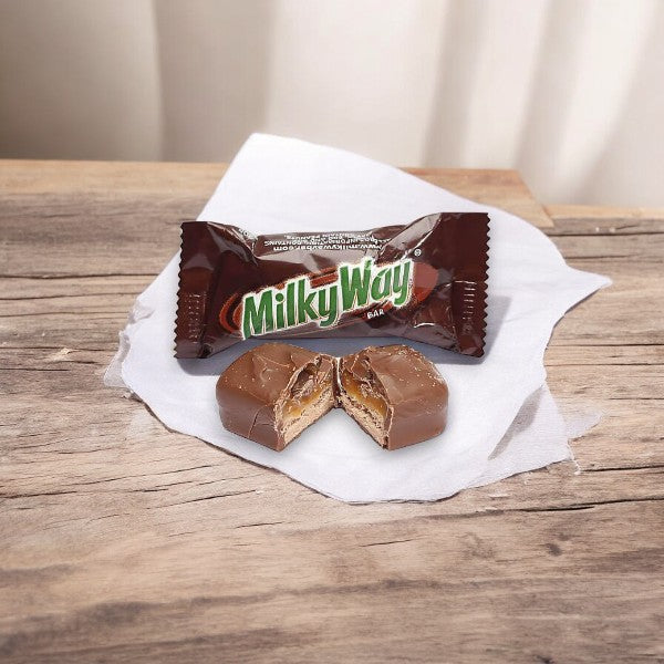 A Fun Size MilkyWay bar from the 5 Count Fun Size Bars pack by Mars, cut in half to show its caramel and nougat filling, is placed on a white napkin on a wooden surface.