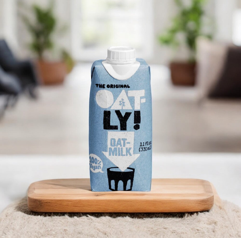 A carton of Oatly Original Oat Milk, the preferred choice for vegans, stands on a wooden tray. The light blue packaging prominently features the text "Oatly Original Oat Milk" and "100% Vegan." The background showcases a slightly blurred indoor setting, making this plant-based beverage an ideal dairy-free alternative.