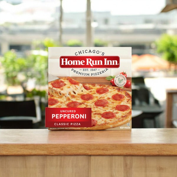 A box of Easy Lunches Home Run Inn Uncured Pepperoni Classic Pizza sits on a wooden table with an outdoor setting in the background, next to a variety pack that includes Sausage Pizza, Sausage & Pepperoni Pizza, Pepperoni Pizza, and Cheese Pizza to satisfy every taste.