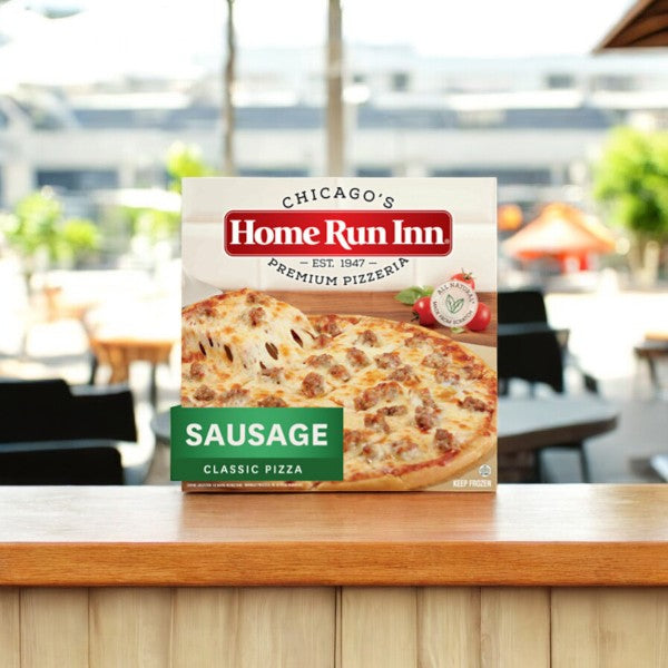 A box of Easy Lunches Home Run Inn Family Sausage Pizza, 30 oz. - 1 Count, with its buttery flaky crust is displayed on a wooden counter, with an outdoor seating area in the background.