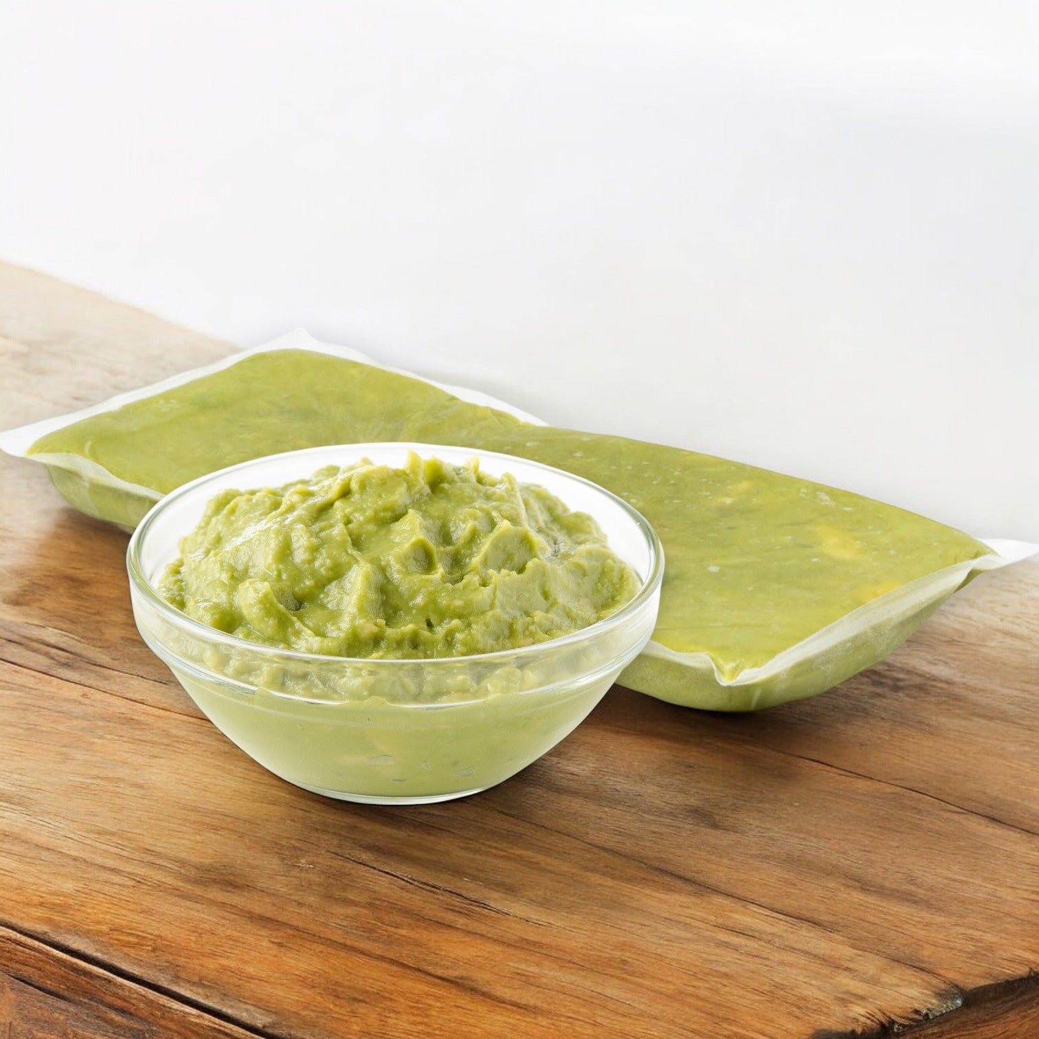 A small glass bowl filled with mashed Hass avocados sits on a wooden surface, with two sealed packs of Wholly Hass Guacamole from Wholly in the background.