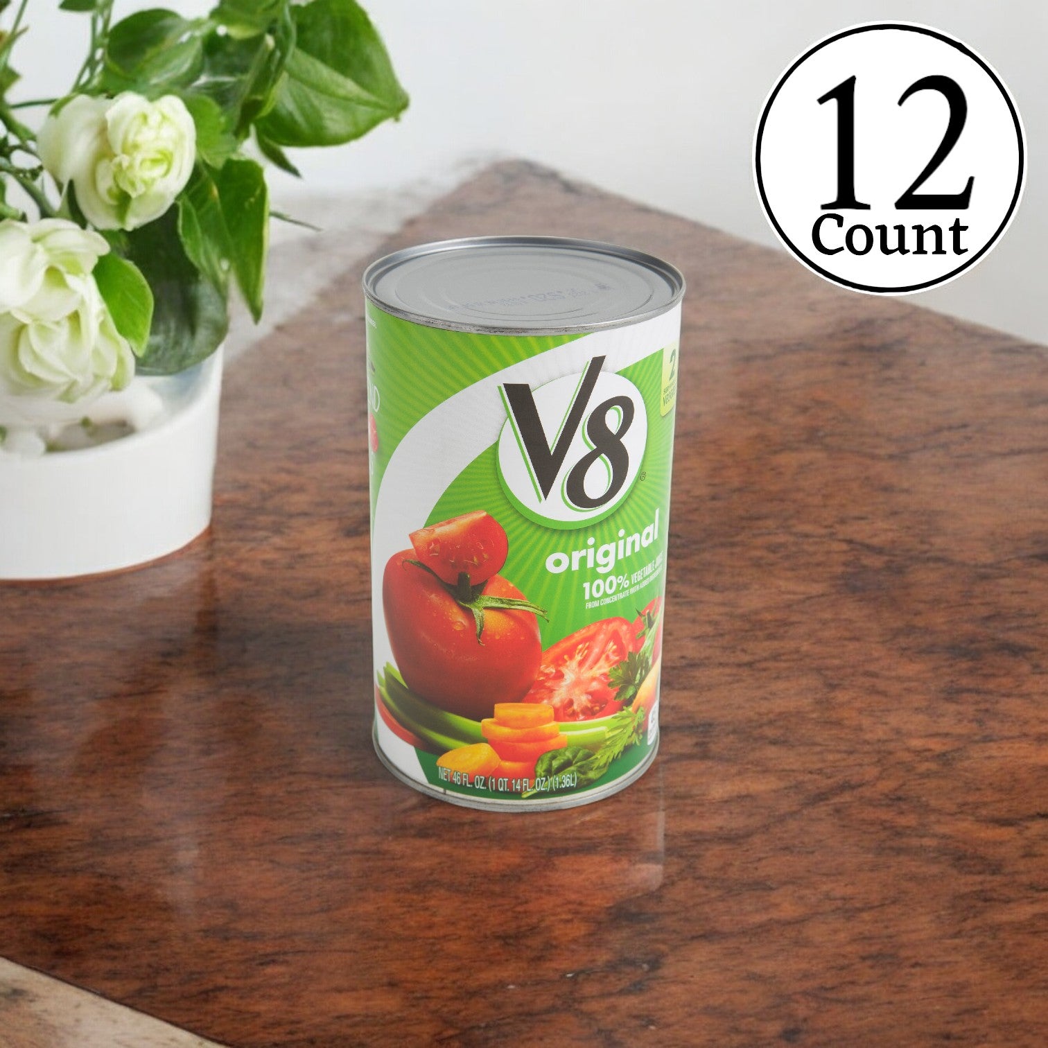 A 46 oz. can of V8 Vegetable Juice from a case of 12 packs sits on a wooden table. In the background, a potted plant and a "12 Pack" label emphasize its rich supply of vitamins and antioxidants.