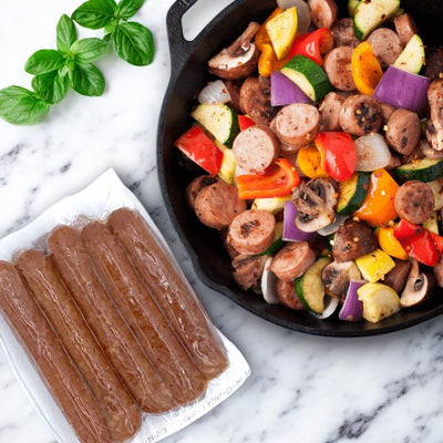 A skillet with sliced chicken sausage, mushrooms, and colorful vegetables sits next to a pack of Amylu Chicken Sausage Links – Apple & Maple on marble. Fresh basil leaves beside them highlight this delightful gluten-free option.