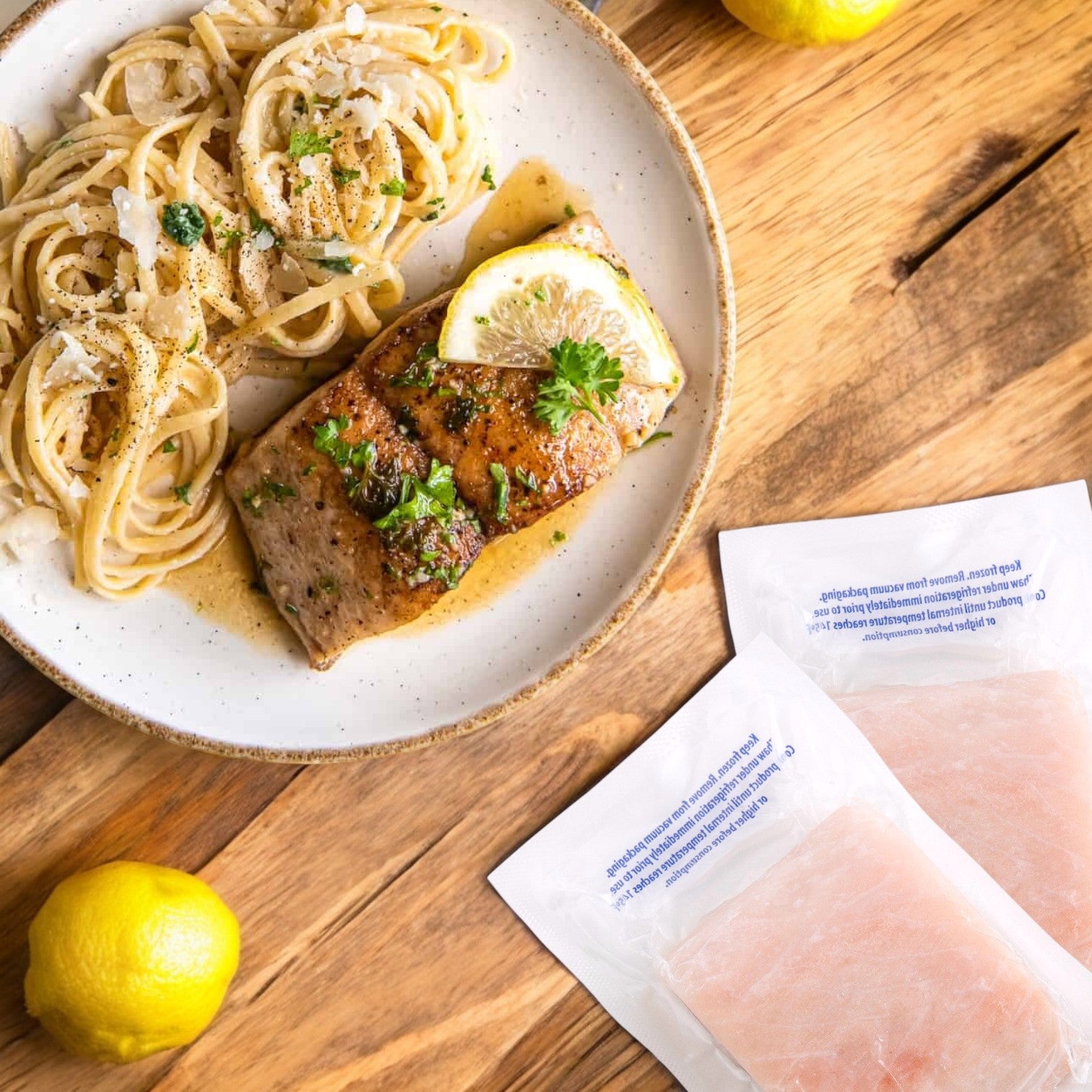 A plate of pasta topped with a slice of wild-caught Mahi Mahi, garnished with lemon and parsley, is surrounded by lemons and packs of Portico Classic Seafood Boneless & Skinless Mahi Mahi Portions (7-9 oz.) on a wooden surface.