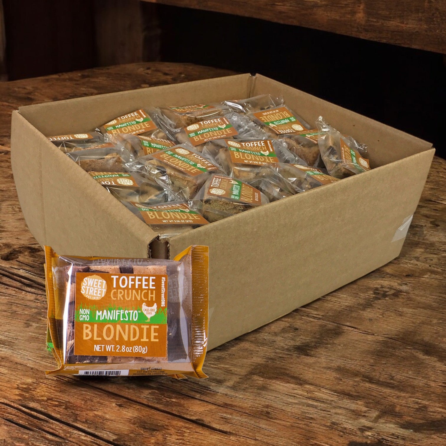 A cardboard box holds Sweet Street Blondie Toffee Crunch Brownies, 48 individually wrapped at 2.8 oz each. One bar is featured upfront, displaying the brand name, weight, and a Non-GMO promise for those seeking premium desserts.