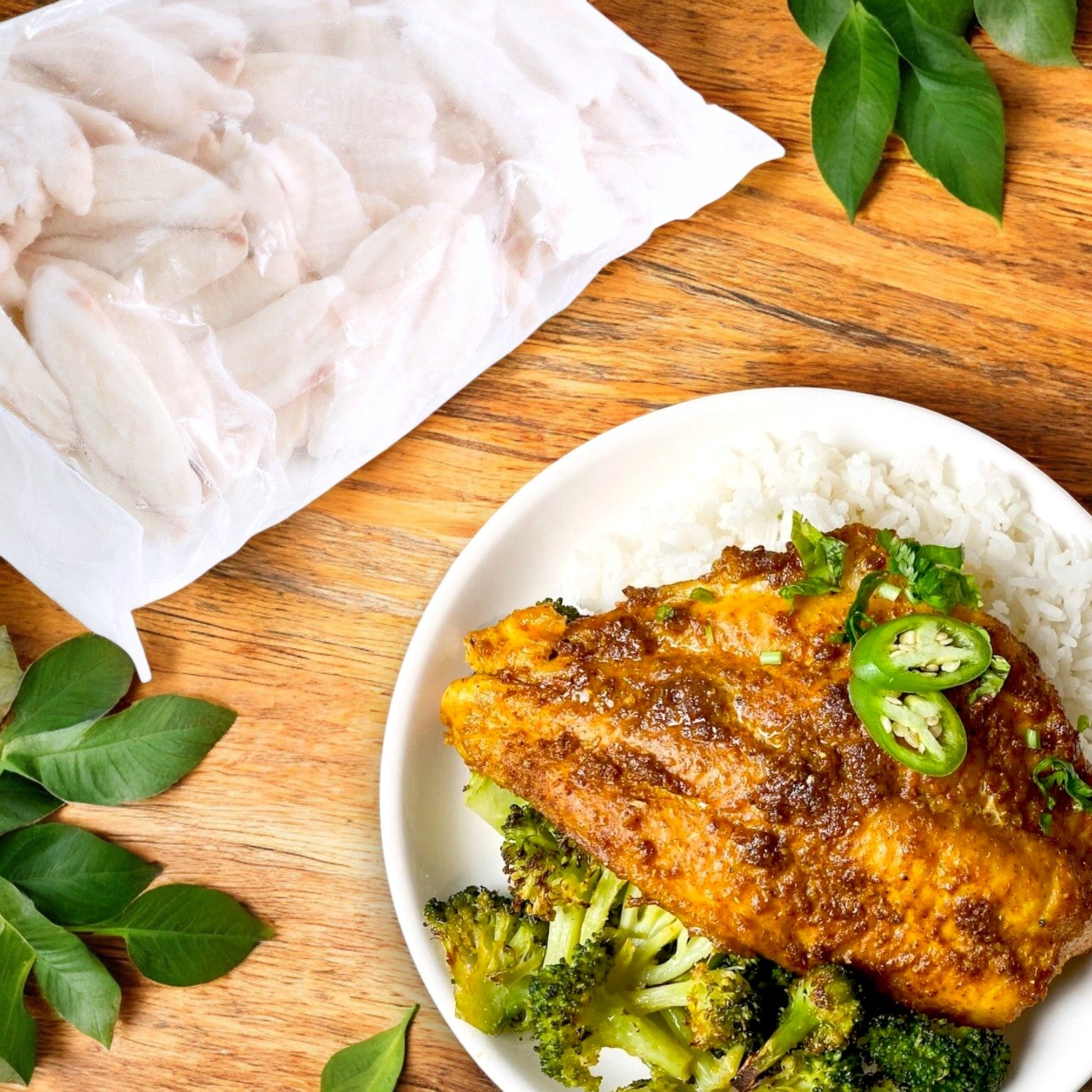 A plate of curry-marinated tilapia fillet with sliced jalapeños, rice, and broccoli sits next to a package of Portico Simply Tilapia Fillets – Boneless & Skinless (IQF, 3-5 oz., 10 lb. Case) on a wooden table adorned with green leaves.