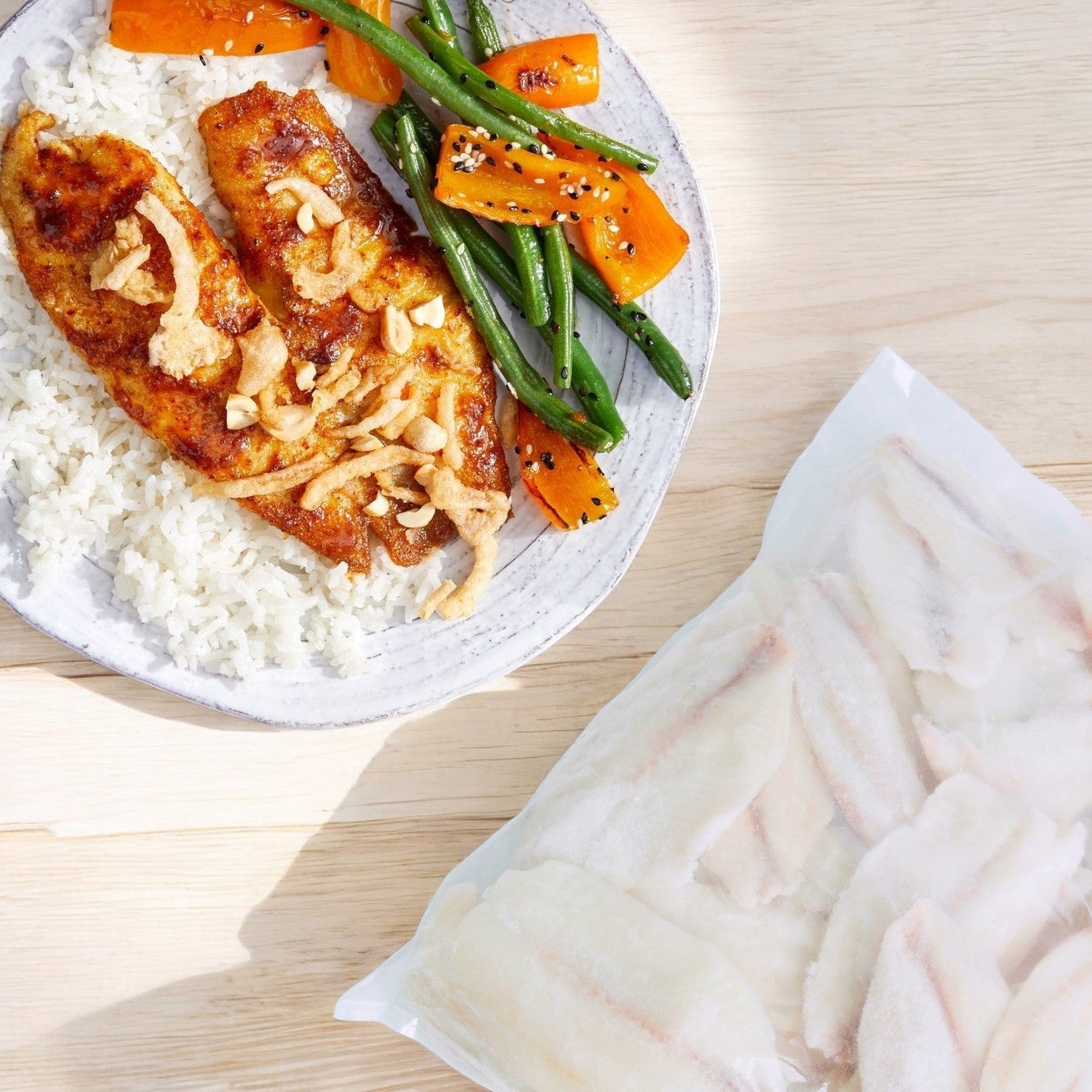 A plate featuring grilled tilapia fillets, rice, carrots, and green beans sits beside a Portico Classic Seafood bag of Portico Simply Tilapia Fillets - Boneless & Skinless (IQF, 5-7 oz., 10 lb. Case) on a wooden surface.