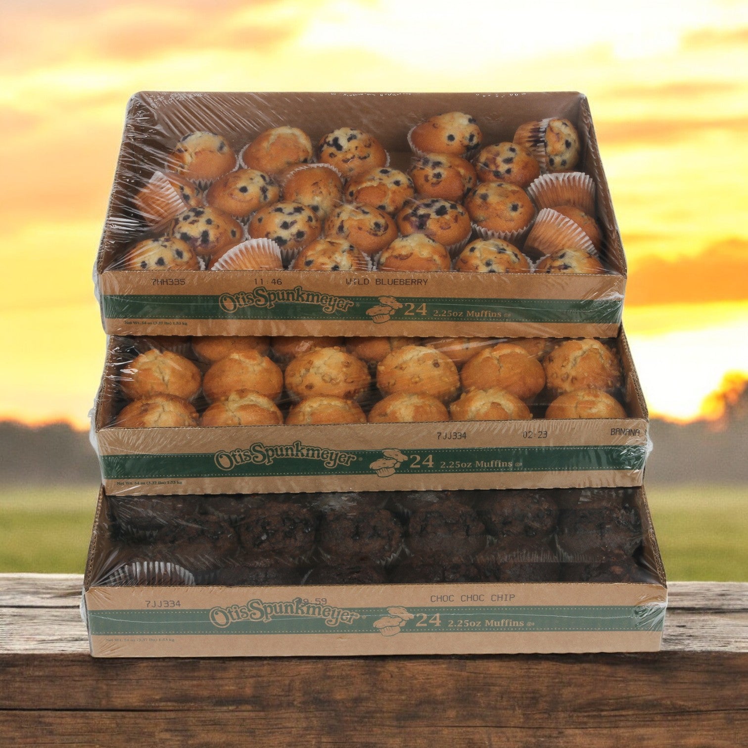 Three open boxes of Otis Spunkmeyer Muffin Variety sit stacked on a wooden table. The bulk packaging reveals the delicious selection: blueberry, banana nut, and chocolate chip muffins. Out of 96 total muffins, the rich aroma of the chocolate chip flavor is particularly tempting.