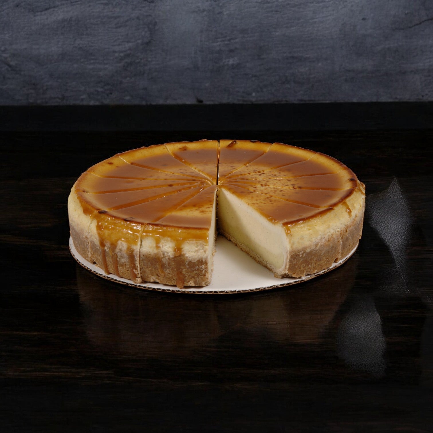 The Sweet Street Cheesecake Creme Brulee, showcased with a slice taken out and elegantly arranged on a white round board against a dark background, boasts an irresistible caramelized sugar crust similar to crème brûlée.