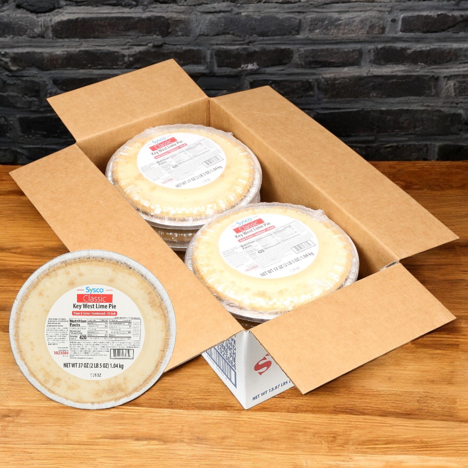 An open cardboard box on a wooden surface displays multiple Sysco Classic Key Lime Pies, their packaging suggesting tropical flavors. One lone pie, highlighting its zesty key lime juice origin, is facing the camera.