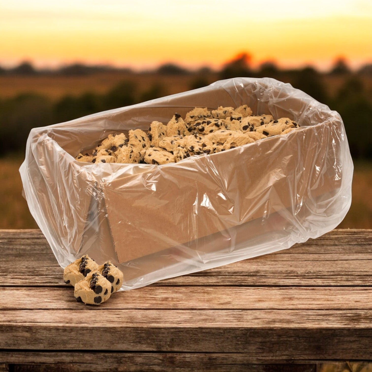 A wooden table with a sunset in the background holds a cardboard box lined with plastic containing David's Cookies Dough Cookie - Chocolate Chip (Raw, 80/4 oz) by David's Cookies.