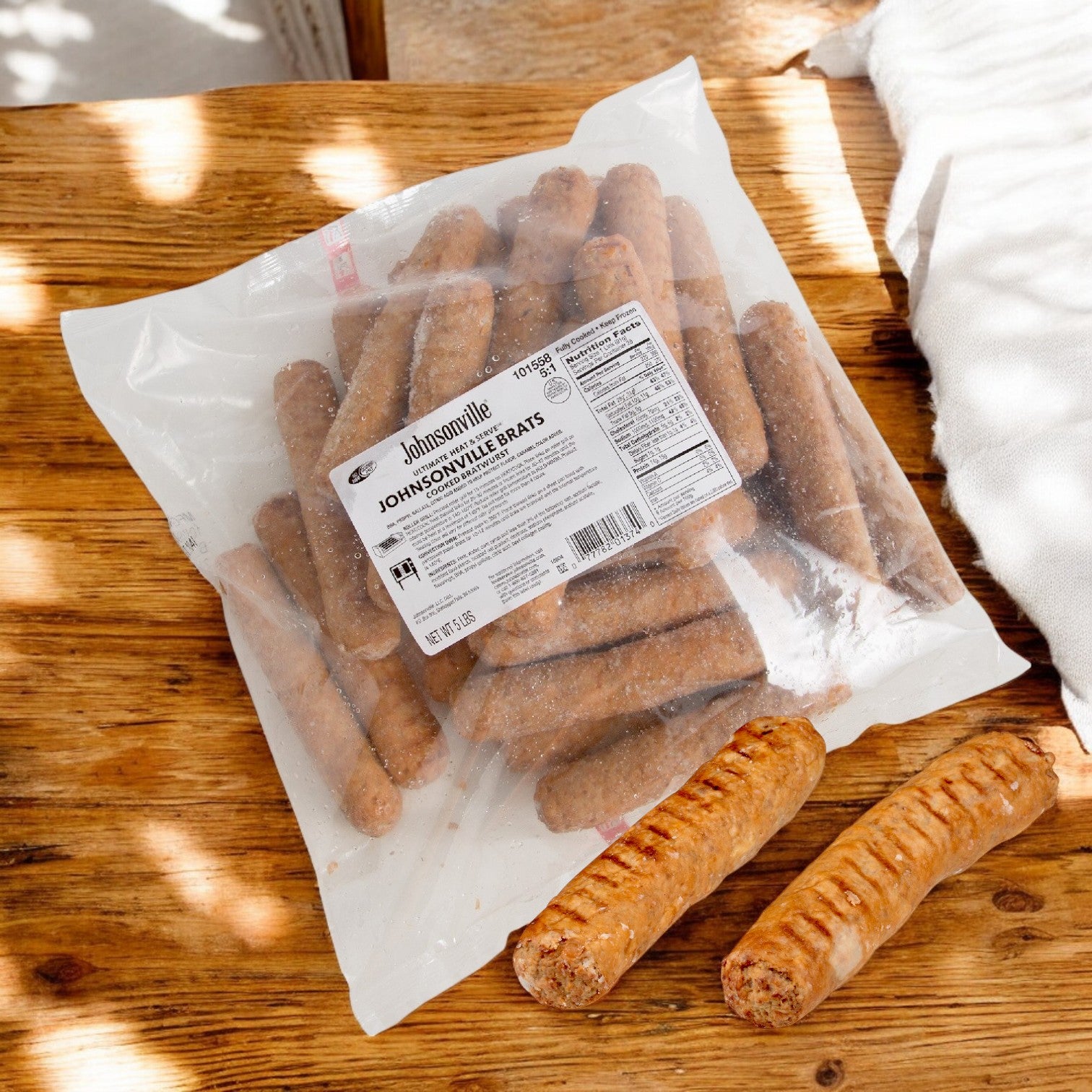 A package of Johnsonville Foods Bratwurst Pre-cooked Ultimate 5x1 (2/5 LB) sits on a wooden table. Beside it, two cooked bratwursts are displayed.