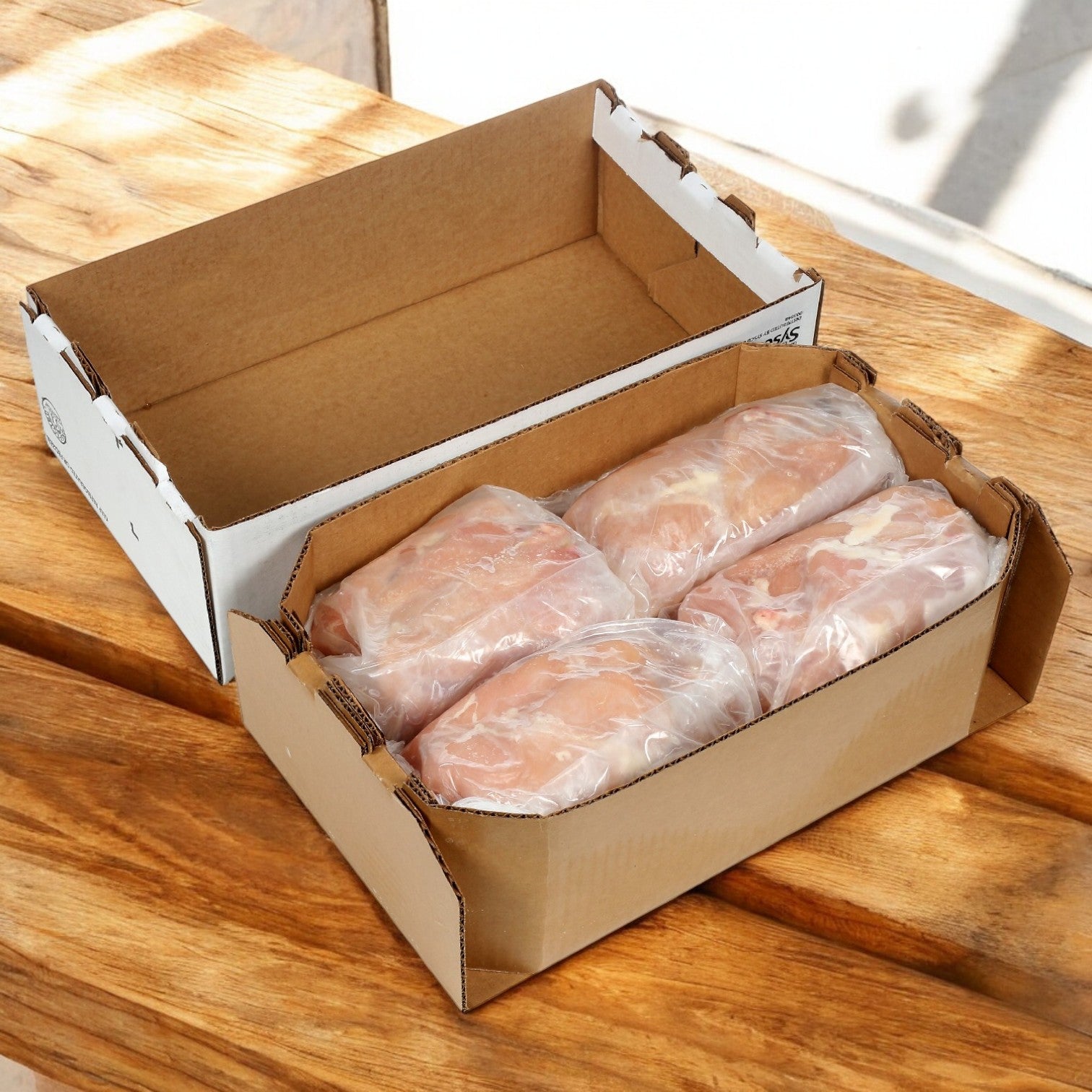 Four vacuum-sealed packs of Sysco Classic Chicken CVP Whole Breast nestle in an open cardboard box on a wooden surface, ready to enhance any foodservice operation with quality and flavor.