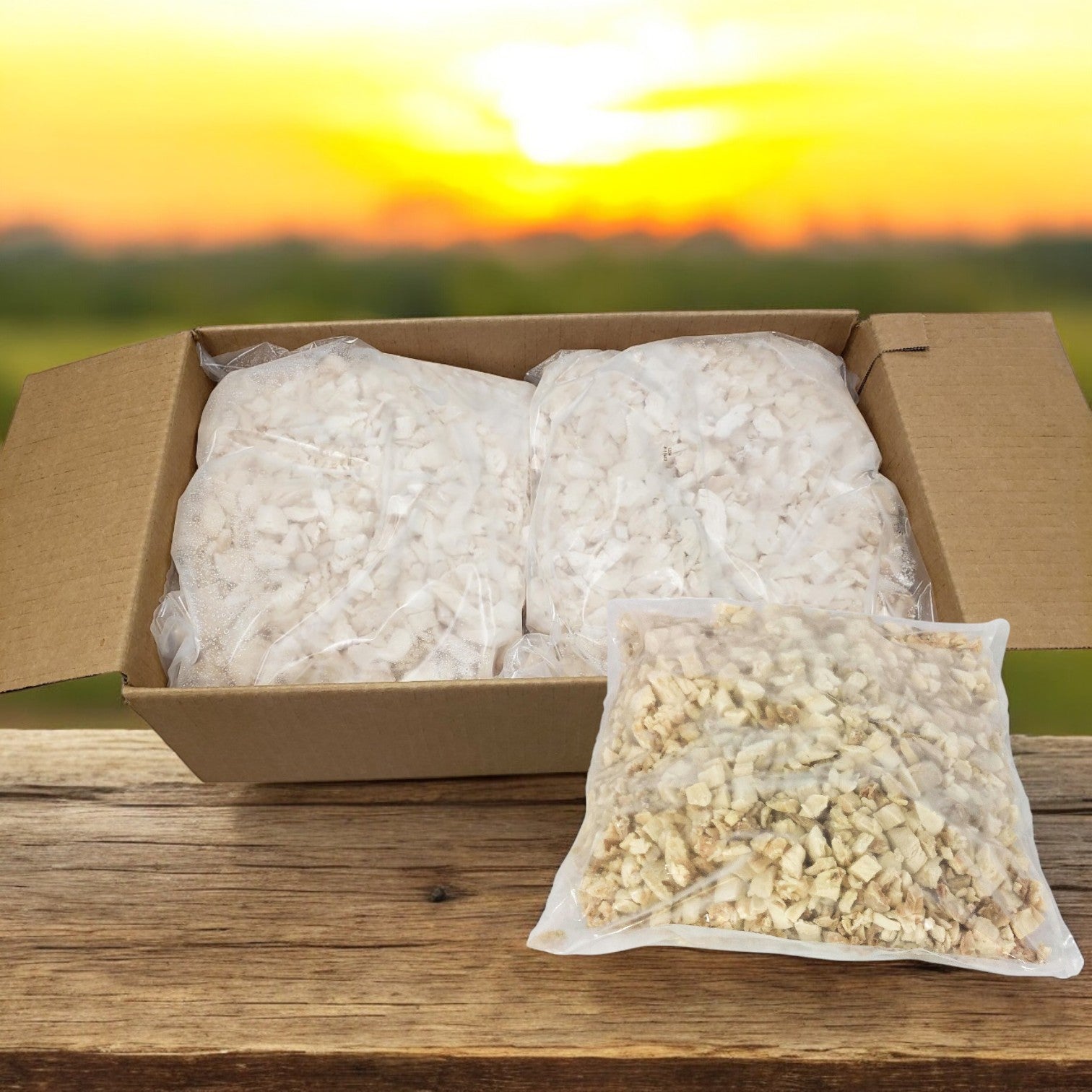 A Sysco Classic box of Diced White Chicken Meat (Low Sodium, 2/5 LB) containing three vacuum-sealed bags rests on a wooden table against a sunset backdrop.