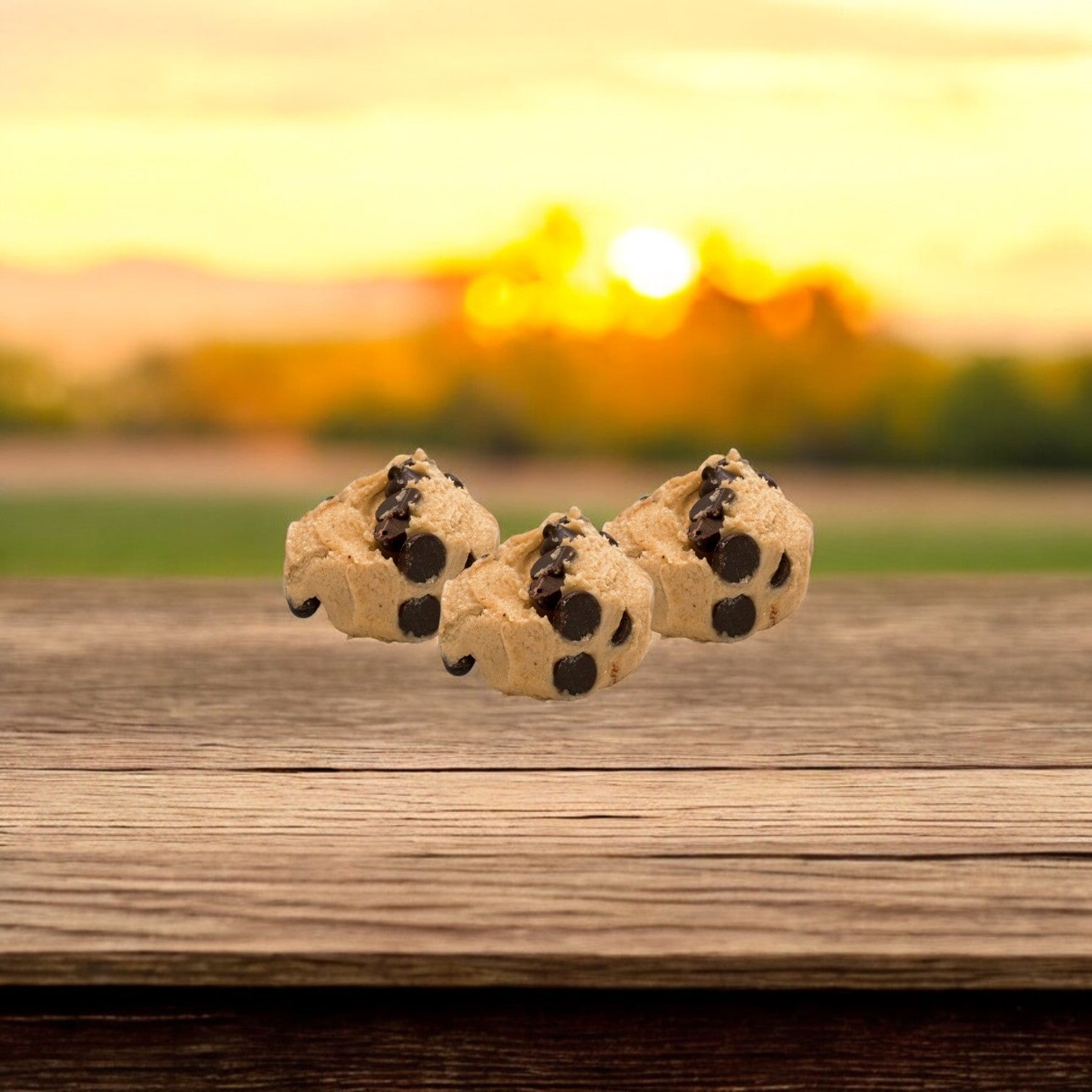 Three freshly baked chocolate chip cookies from David's Cookies Dough Cookie - Chocolate Chip (Raw, 80/4 oz) rest on a wooden surface, bathed in the warm hues of a sunset.