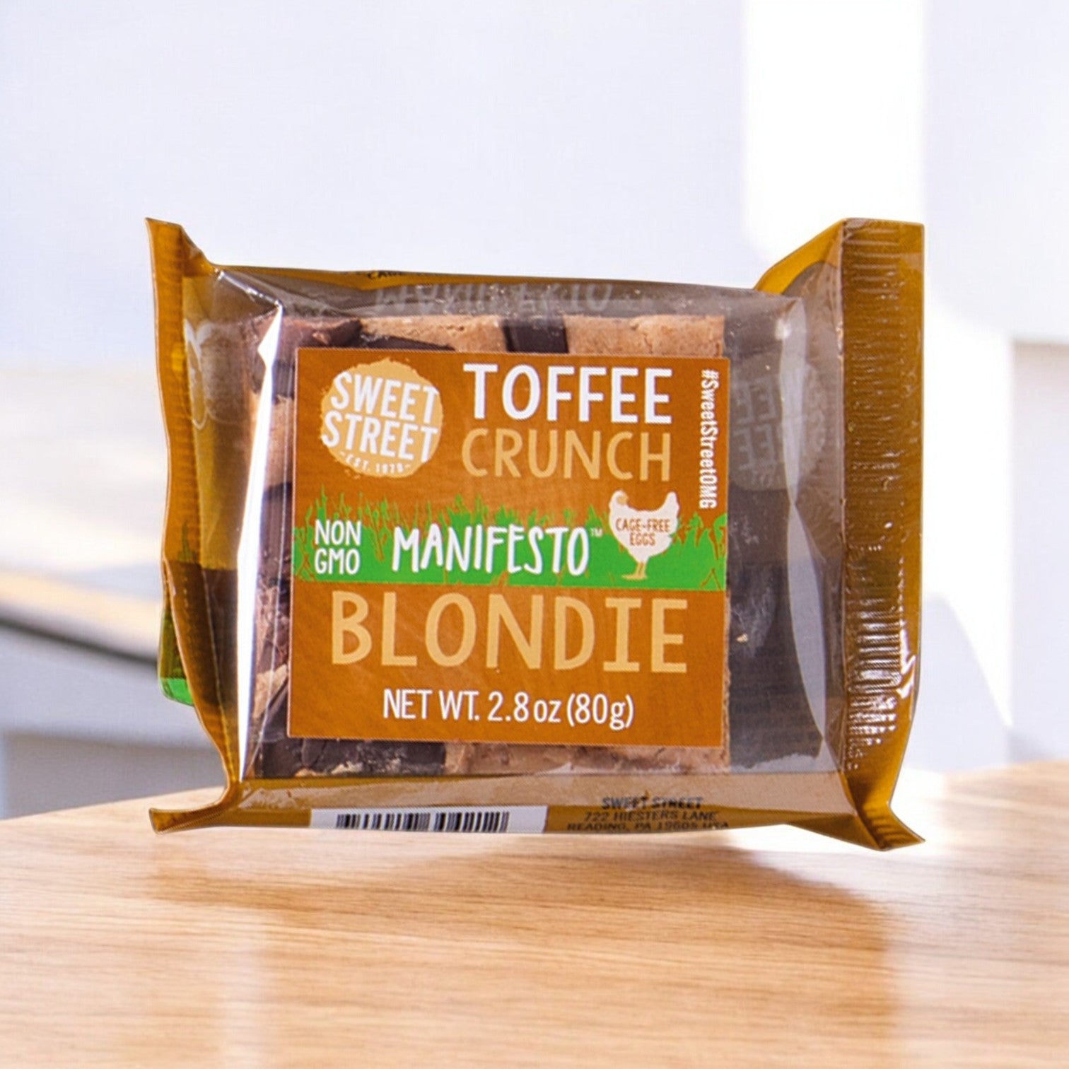 A 2.8 oz. Sweet Street Blondie Toffee Crunch Brownie from a 48-count pack sits on wood, featuring non-GMO ingredients and cage-free eggs for a premium dessert experience.