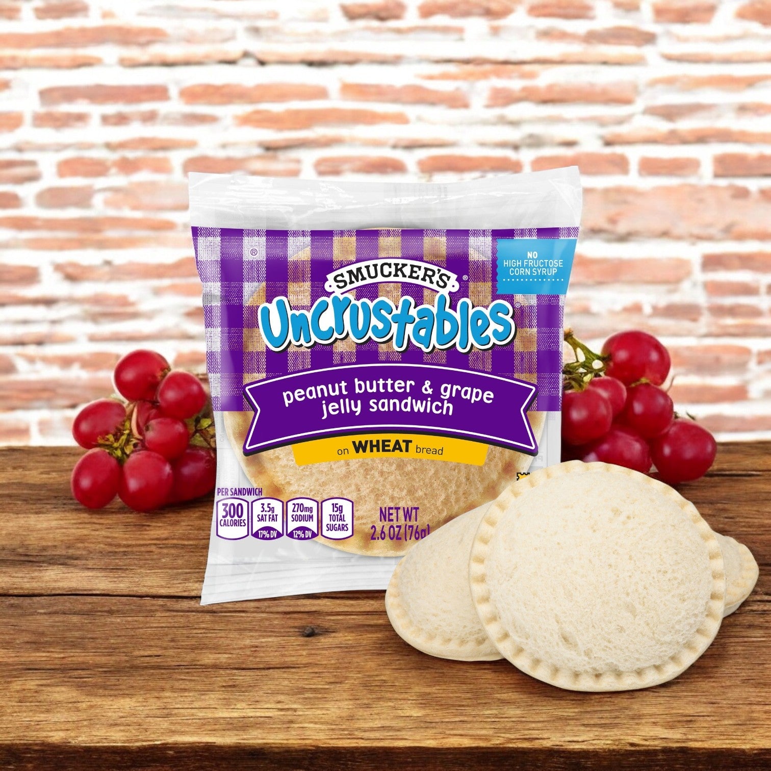 Uncrustable Smuckers Peanut Butter & Grape Jelly Wheat 2.6 oz. Sandwiches, available in a pack of 48, are placed on a wooden surface with vibrant red grapes in the background.