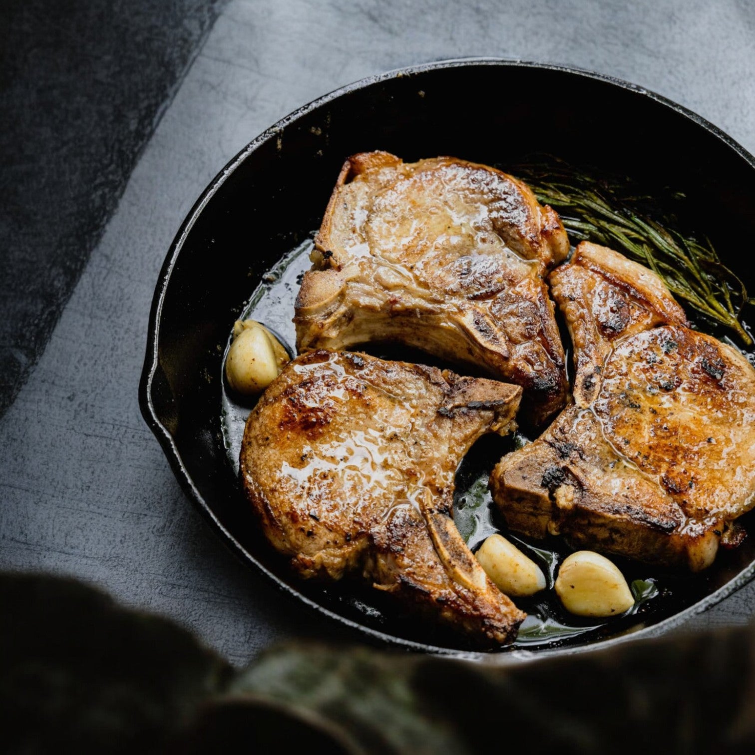 Three Buckhead Pride/Newport Pride bone-in center-cut rib pork chops sizzle in a cast iron skillet, surrounded by aromatic garlic cloves and fragrant rosemary sprigs.