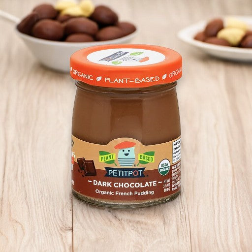 A jar of Petit Pot's Organic Oatmilk Chocolate French Dessert rests on a wooden surface, while two bowls with assorted nuts remain blurred in the background.