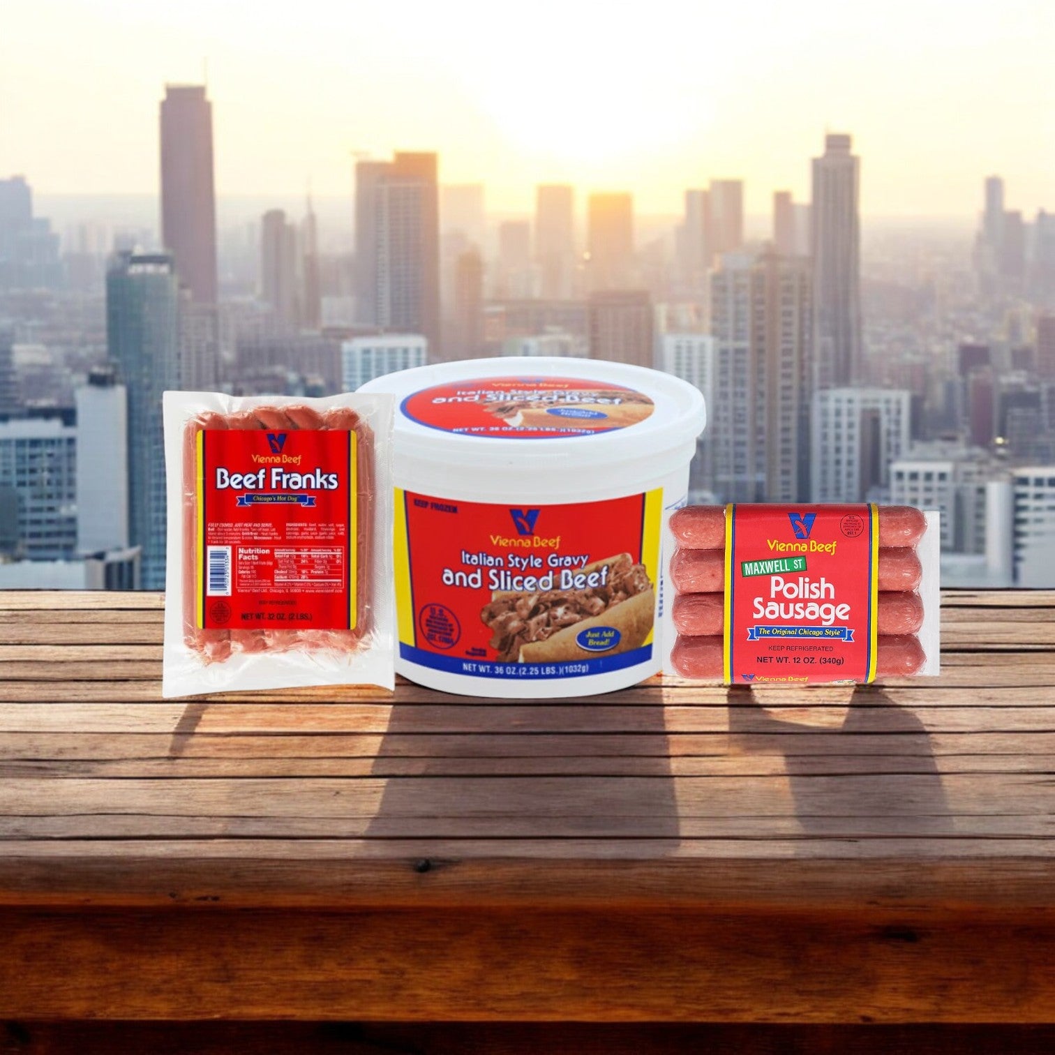 Packages of Easy Lunches' Chicago Classic Meat Combo, featuring fully cooked franks, Italian beef, and Maxwell St. Polish Sausage, are beautifully arranged on a wooden table with a city skyline in the background.