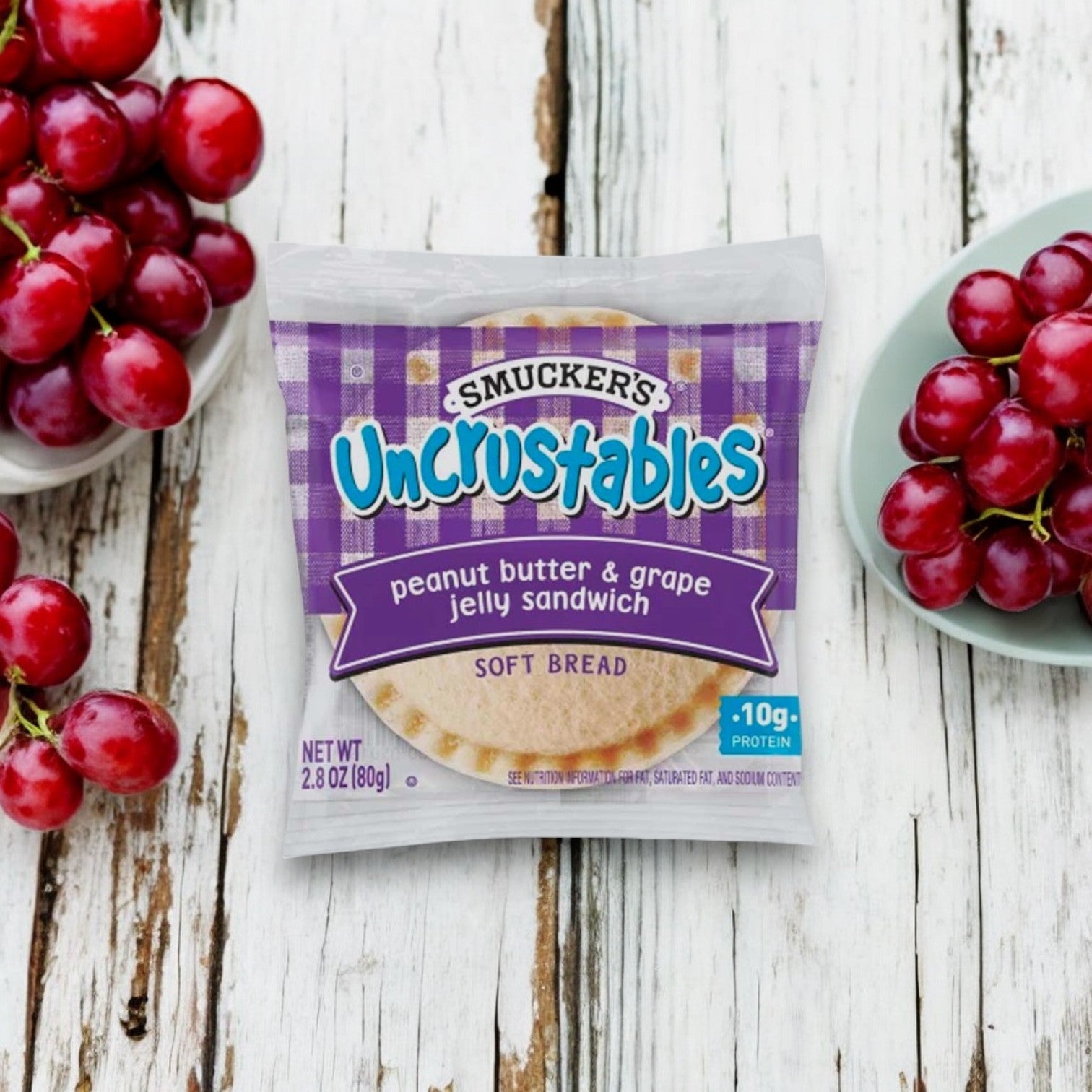 An Uncrustables Peanut Butter & Grape Jelly Sandwich 1 Sandwich by Uncrustable Smuckers is displayed on a white wooden surface, flanked by two bowls of fresh red grapes. Perfect for a quick snack!