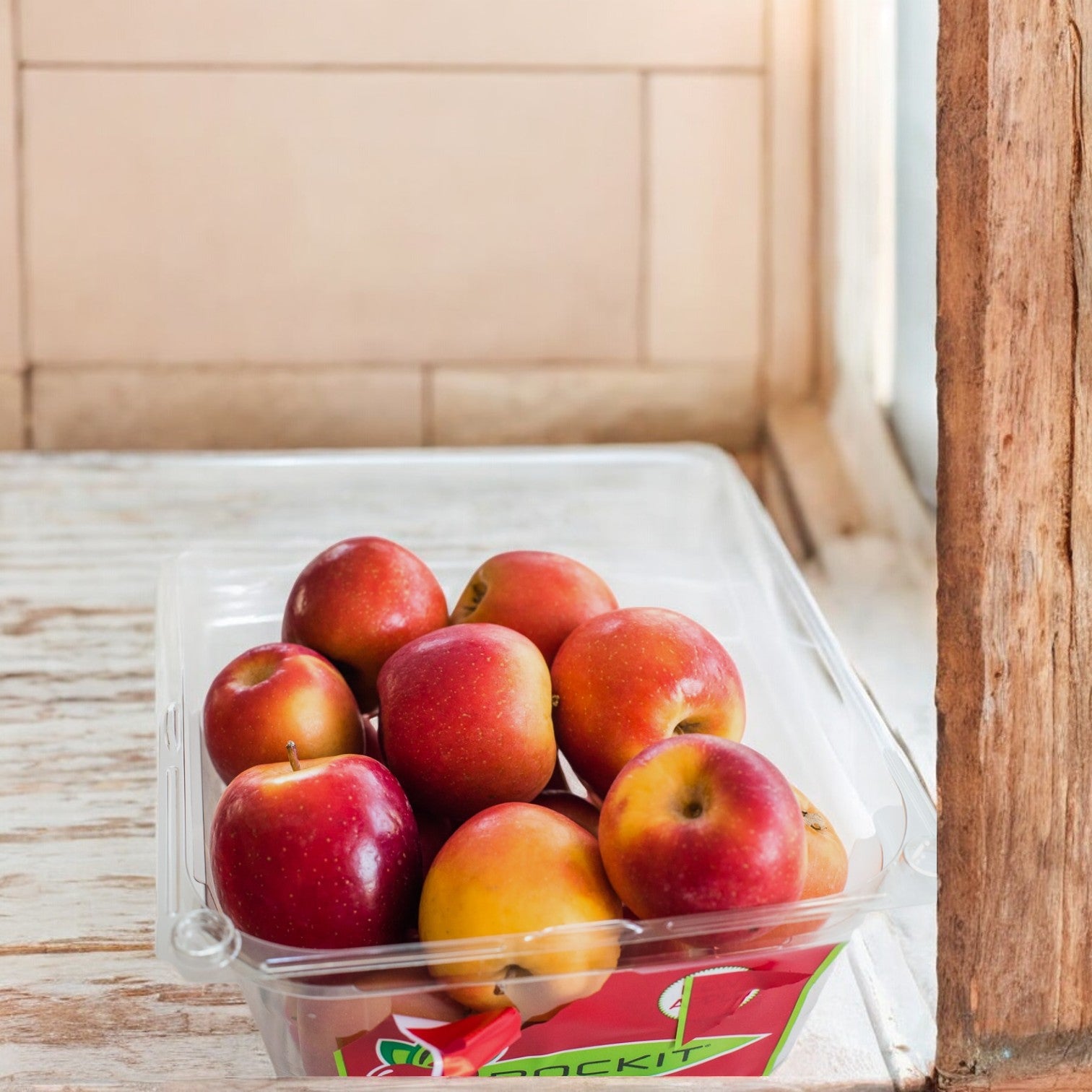 A wooden surface near a window holds a plastic container filled with Easy Lunches' Rockit Crisp Sweet Miniature Apples.